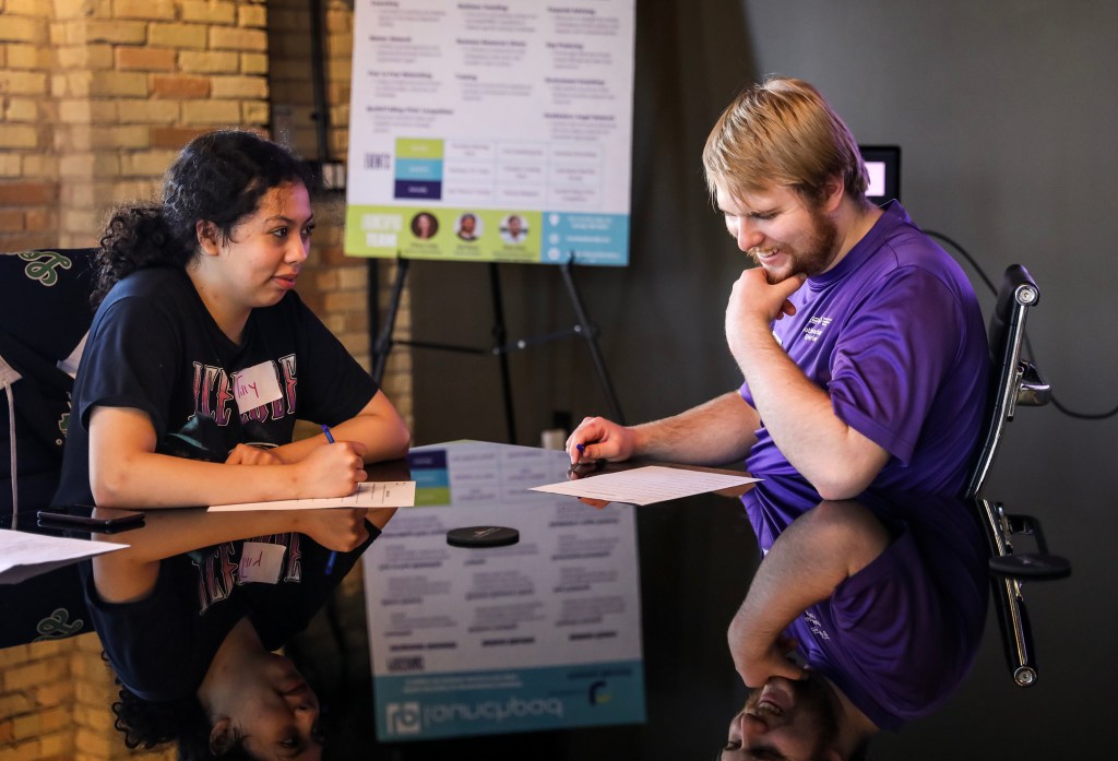 Two youth sit at a board room table smiling. On the left is a young native woman with light brown skin and dark brown curly hair. She is wearing a black t-shirt. On the right is a young white man with short blond hair wearing a purple t-shirt.
