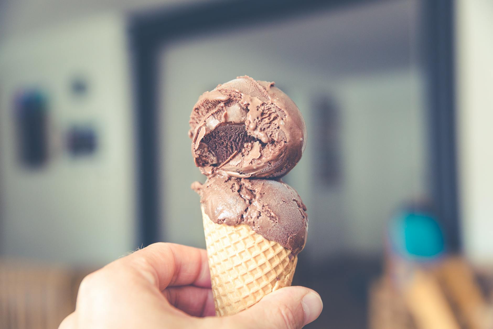A hand holds out a chocolate ice cream cone with a blurred background