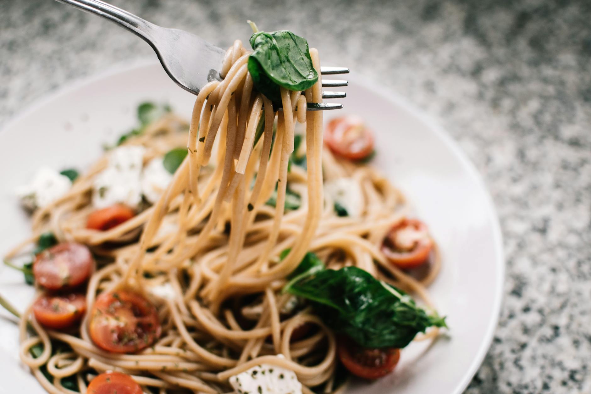 Pasta dish with basil, mozzarella and cherry tomato halves on a white plate with a granite counter in the background. A fork is scooping up some of the pasta.