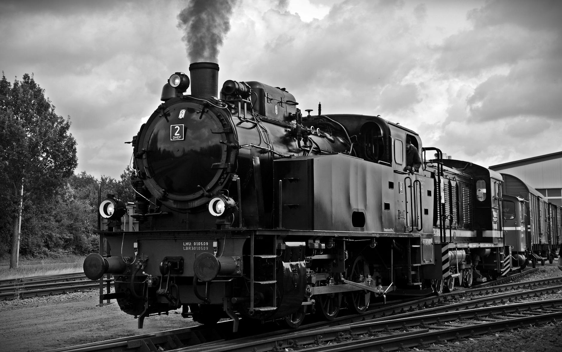 Close up black and white image of a train engine with more train cars behind it. There is smoke coming out of the smokestack.