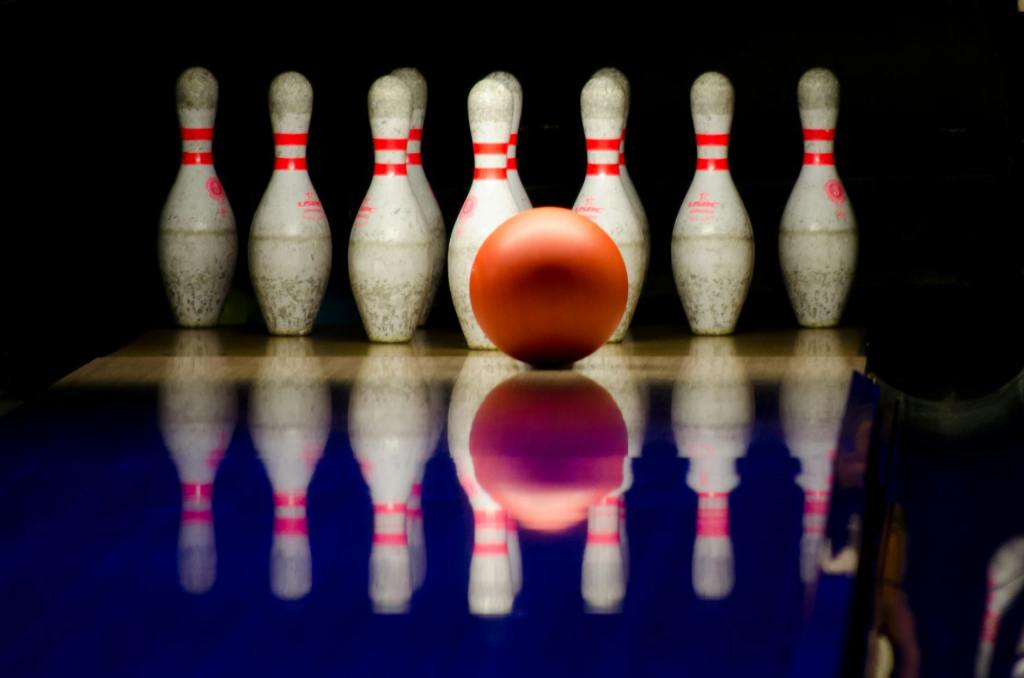 Bowling pins on a dark alley lane. An orange ball is in front of the pins and there is a reflection of the balls and pins on the lane.